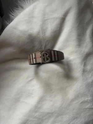 Rutig Burberry bälte med B-spänne - Säljer ett snyggt Burberry bälte med klassiskt rutig design i beige, svart och röd. Bältet har ett metallspänne med ett stort B framtill. Materialet är slitstark syntet med läder på insidan. Perfekt för att lyfta din outfit med en lyxig touch. Väldigt sparsamt använt. Storlek 80. Kom med bud 