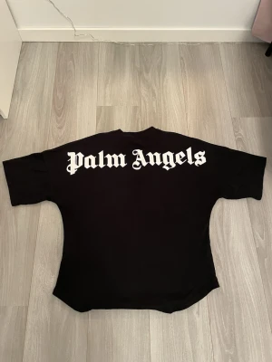 Svart Palm Angels t-shirt oversized - Svart oversized t-shirt från Palm Angels med stort vitt tryck på ryggen och mindre logga vid halsen. Klassisk rund halsringning och korta ärmar. Tillverkad i mjuk bomull för skön känsla och streetwear-vibe.