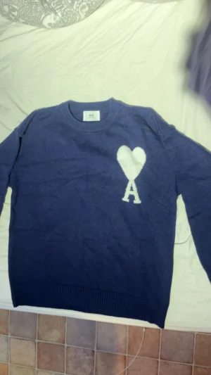 Blå stickad tröja från Ami - Säljer en mörkblå stickad tröja från Ami Alexandre Mattiussi med ett vitt hjärta och bokstaven A på bröstet. Tröjan har rund hals, ribbade muddar och lång ärm. Perfekt för dig som gillar stilrena detaljer och vill ha något som sticker ut lite extra.