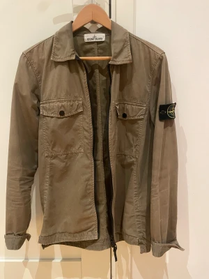 Olive green overshirt från Stone Island - Säljer en olivgrön overshirt från Stone Island med klassisk patch på ärmen. Jackan har två bröstfickor med knappar, dragkedja framtill och en stilren krage. Perfekt för lager-på-lager och snygg till jeans eller cargo.
