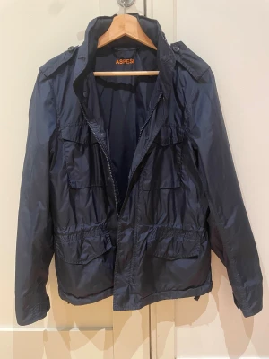 Mörkblå vindjacka från Aspesi - Snygg mörkblå vindjacka från Aspesi med flera praktiska fickor framtill och dragkedja. Jackan har en klassisk krage, långärmade ärmar och är tillverkad i lätt polyester som passar perfekt för blåsiga dagar. Stilren design med diskreta detaljer.