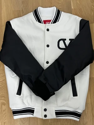 Vit och svart bomberjacka från Suprem - Snygg bomberjacka från Suprem i vit med svarta ärmar och detaljer. Jackan har ribbade muddar med svarta och vita ränder, knappar framtill och ett stort tryckt emblem på bröstet. Materialet är vattenavvisande och känns riktigt fräscht. Perfekt för dig som vill sticka ut.