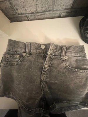Grå denimshorts från 157 - Snygga grå denimshorts från 157 med markerade vita sömmar och klassisk femficksdesign. Modellen är högmidjad och har knappar och dragkedja framtill samt fickor med lock och knapp bak. Perfekta för en chill och avslappnad stil.