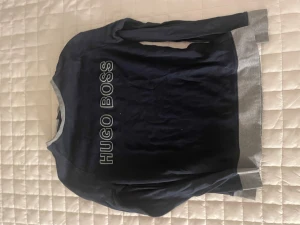 Mörkblå tröja från Hugo Boss - Snygg mörkblå långärmad tröja från Hugo Boss med gråa muddar vid hals, ärmslut och nederkant. Stor logotyp framtill i vitt. Perfekt för dig som gillar stilrena märkeskläder och vill ha en enkel men cool look.