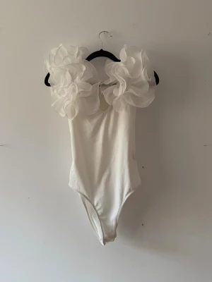 Vit body med volangärmar - Säljer en vit body med stora, fluffiga volangärmar i transparent tyg. Kroppsdelen är slät och enkel, medan axlarna har riktigt statement volanger som ger en cool och unik look. Perfekt för dig som vill sticka ut och älskar trendiga detaljer.