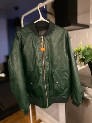 Grön skinn bomberjacka från Prada - Snygg mörkgrön bomberjacka från Prada i skinn med silverfärgad dragkedja och ribbade muddar. Jackan har praktiska fickor med tryckknappar och en unik ficka med Prada-logga på ärmen. Perfekt för dig som vill sticka ut med exklusiv stil.