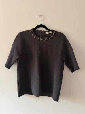 Mörkgrå topp med guldkedja från Zara - Snygg mörkgrå topp från Zara med korta ärmar och rund halsringning dekorerad med guldfärgad kedja. Toppen har en rak passform och är tillverkad i ett mjukt material som ger en stilren look. Perfekt att styla med jeans eller kjol för en cool vibe. Ens ringen har lossnat så den hänger lite lös annars är den helt orörd.