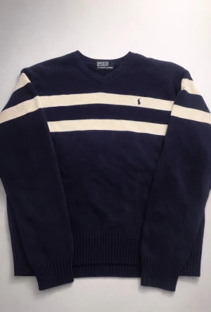 Randig blå tröja från Polo Ralph Lauren - Snygg mörkblå stickad tröja från Polo Ralph Lauren med två breda vita ränder över bröstet och armarna. Tröjan har v-ringad hals och ribbade muddar vid ärmslut och nederkant. Klassisk logga broderad på bröstet. Perfekt för dig som gillar stilren design.