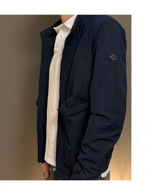 Marinblå vindjacka från J.Lindeberg - Snygg marinblå vindjacka från J.Lindeberg i storlek M. Jackan har klassisk krage, dragkedja framtill, två stora fickor med lock och ribbade muddar vid ärmslut och nederkant. Perfekt för dig som gillar stilren och sportig look.