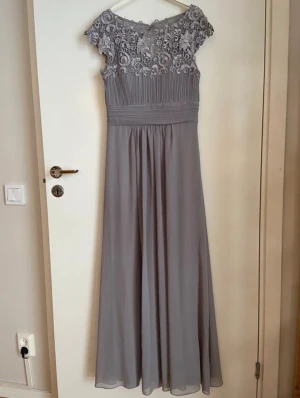 Grå långklänning med spets - Säljer en elegant grålila långklänning med vacker spets upptill och plisserad midja. Klänningen har ärmlös design och detaljerad broderad spets över axlar och bröst. Kjolen är mjukt fallande och ger en romantisk look. Perfekt för bal, bröllop etc. Använd en gång som brudtärneklänning. Uppsydd hos sömmerska på längd och axlar. Jag är 160cm lång och den passar perfekt i längden med 7 cm klack. 