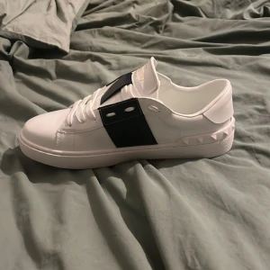 Valentino Garavani vita sneakers - Snygga vita sneakers från Valentino Garavani med bred svart rem över snörningen och coola nitar längs hälen. Skorna har platt sula och rund tå, tillverkade i skinn med klassisk siluett och diskret logga på sulan. Perfekt för dig som gillar stilrena designer med edge.