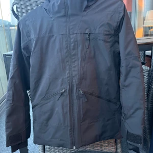 Svart jacka från Everest stl. 152 - Svart jacka från Everest med huva och flera praktiska fickor med dragkedja. Jackan har justerbara ärmslut med kardborre och snygga logodetaljer på både bröstet och huvan. Perfekt för snön och skidåkning.