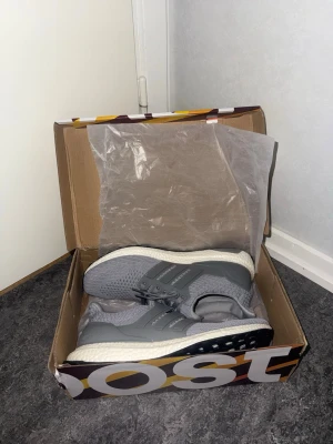 Adidas Ultraboost grå sneakers - Säljer ett par grå Adidas Ultraboost sneakers med stickad ovandel och snörning. Skorna har en vit och svart platt sula med boost-teknologi för extra komfort. Perfekta för dig som gillar stilrena och sportiga skor med modern känsla.