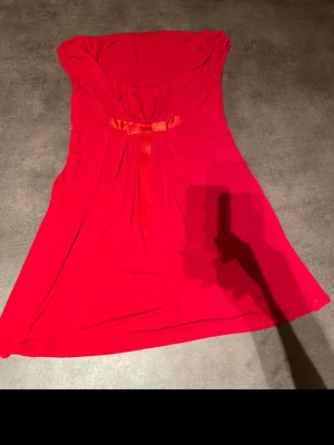 Röd kortklänning med rosett - Säljer en röd kortklänning med söt rosett framtill och lätt puffade axlar. Klänningen har en mjuk och flowy passform, rundad urringning och är tillverkad i ett tunt, skönt material som känns bekvämt mot huden. Perfekt för dig som gillar färgstarka och lekfulla plagg.