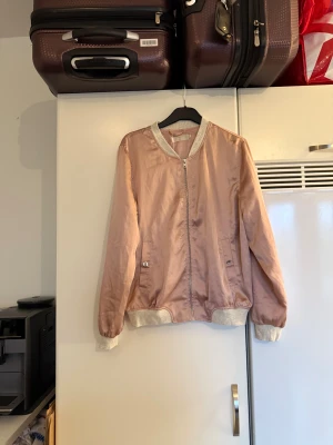 Rosa bomberjacka från Cream - Snygg rosa bomberjacka från Cream med grå ribbade muddar och dragkedja framtill. Jackan har coolt tryck med blommor och siffror på ryggen, samt fickor med knappdetalj. Materialet är glansigt och känns lätt på kroppen. Perfekt för dig som vill sticka ut lite extra. Storlek M/38. Fin men tyvärr kan tråddraningar finnas. 