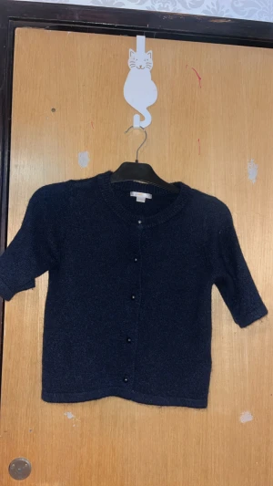 Mörkblå stickad kofta från Gina Tricot - Snygg mörkblå stickad kofta från Gina Tricot med rund hals och korta ärmar. Koftan har pärlknappar framtill och ribbade kanter vid hals och ärmslut. Perfekt för lager-på-lager och chill stil. Kan va stickig