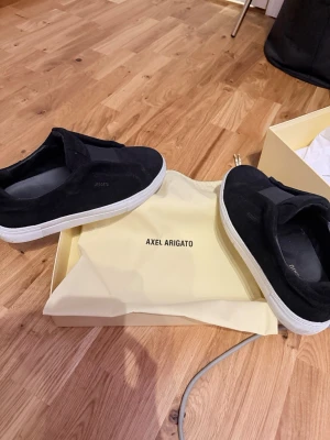 Svarta sneakers från Axel Arigato - Svarta sneakers från Axel Arigato med stilren design och vit platt sula. Skorna är tillverkade i mocka och har en låg siluett utan snörning, vilket ger en clean och modern look. Perfekta för dig som gillar minimalistisk stil och premiumkänsla.