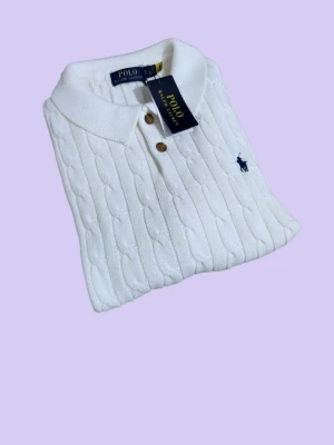 Vit stickad pikétröja från Polo Ralph Lauren - Vit stickad pikétröja från Polo Ralph Lauren med klassisk kabelstickad struktur, krage och bruna knappar framtill. Tröjan har korta ärmar och den ikoniska broderade loggan i marinblått på bröstet. Perfekt för en clean och stilren look.