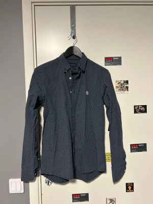 Mörkblå prickig skjorta Henri Lloyd - Snygg mörkblå skjorta från Henri Lloyd med små vita prickar och klassisk krage. Skjortan har långa ärmar, vita knappar och ett broderat emblem på bröstet. Perfekt för dig som gillar stilrena detaljer och bekvämt material.