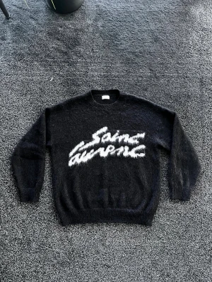 Saint Laurent stickad tröja - Säljer en svart stickad tröja från Saint Laurent med vit, fluffig logotyp på bröstet. Tröjan har rund hals, långa ärmar och ribbade muddar. Materialet är mjukt och har en lite lurvig yta som ger en cool vibe. Perfekt för dig som gillar exklusiva märken och streetstyle. Skick 10/10, storlek M men passar även L. Pris går att diskutera✅