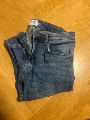 Blå jeans från 157 med slim fit - Snygga blå jeans från 157 med klassisk femficksdesign och smal passform. Jeansen har en mörkblå tvätt med lätt slitning och bruna sömmar. Märkespatch baktill i brunt. Perfekta för dig som gillar en enkel och stilren look.