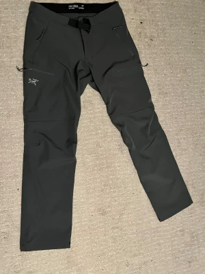 Svarta Arc'teryx cargopants med bälte - Arc'teryx cargopants i svart med raka ben, inbyggt bälte och flera dragkedjeförsedda fickor. Byxorna har stilren design med Arc'teryx-logga på benet och är gjorda i slitstark syntet. Perfekta för utomhusaktiviteter och äventyr.