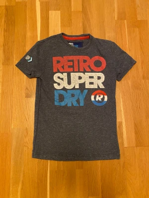 Retro Superdry t-shirt mörkgrå - Snygg mörkgrå t-shirt från Superdry Retro Sport med tryck i rött, vitt och blått på framsidan. Klassisk rund hals och korta ärmar. Materialet är mjuk bomull och har en melerad look. Märkespatch på ärmen för extra detalj. Perfekt för dig som gillar streetwear.