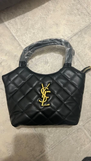 Svart quiltad YSL handväska - Säljer en svart quiltad handväska från YSL med guldigt logodetalj framtill. Väskan har en stilren och klassisk design med rutmönster och dragkedja upptill. Perfekt storlek för det viktigaste och enkel att bära i handtaget.