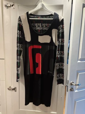 Desigual långärmad mönstrad klänning - Cool långärmad klänning från Desigual med grafiskt mönster i svart, rött, vitt och grönt. Klänningen har texten 'YES' tryckt över hela ärmarna och en stor röd detalj framtill. Materialet är mjukt och stretchigt, perfekt för dig som gillar unika prints och färgkombinationer. Nedre delen är stickad. 