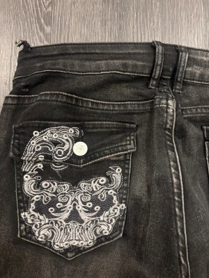 Svarta jeans med broderade fickor - Snygga svarta jeans med vita broderade detaljer på bakfickorna. Jeansen har bootcut-snitt och klassisk femficksdesign. Materialet är jeans och färgen är mörk med lätt tvättad look. Perfekt för dig som gillar unika detaljer och vill sticka ut.
