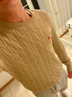 Beige kabelstickad tröja Morris - Säljer en beige kabelstickad tröja från Morris med rund hals och lång ärm. Tröjan har ett litet rött broderat logomärke på bröstet och är gjord i mjuk bomull. Perfekt för dig som gillar klassisk och stilren look.