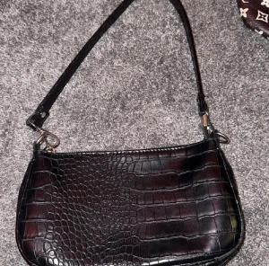 Svart axelväska med krokodilmönster - Snygg svart axelväska med croco-textur som ger en cool och lyxig vibe. Väskan har en avtagbar axelrem och är perfekt för att bära dina essentials. Materialet är skinnimitation med ett glansigt finish och mönstret är inspirerat av krokodil.