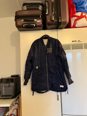 Mörkblå bomberjacka med patches - Snygg mörkblå bomberjacka med broderade patches, bland annat ett stort M på bröstet och detaljer på ärmen. Jackan har dragkedja framtill, ribbade muddar och flera fickor. Perfekt för dig som vill ha en sportig och trendig look. Från märket Odd Molly. Storlek 2 som enligt deras storleksguide motsvarar en M. Mycket fint skick, enda lilla är att knapparna har tappat sin färg. 