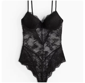 Svart spetsig body med v-ringad urringning - Säljer en svart body i genomskinlig spets med v-ringad urringning och tunna axelband. Bodyn har fina spetsdetaljer och en feminin siluett som ger en snygg look. Perfekt för dig som gillar att sticka ut med stilrena detaljer.