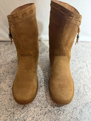 Beige mockaboots från UGG med pärldetalj - Säljer ett par snygga beige boots från UGG i mjuk mocka. Skorna har rund tå, platt sula och dekorativa pärlor och snörning upptill. Snyggt flätad kant runt skaftet och UGG-logga bak på hälen. Perfekt för dig som gillar chill och stilren look.