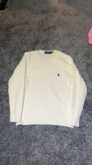 Vit stickad tröja från Polo Ralph Lauren - Snygg vit stickad tröja från Polo Ralph Lauren i storlek S. Tröjan har klassiskt kabelstickat mönster och en broderad marinblå logga på bröstet. Rund hals och ribbade muddar vid ärmslut och nederkant. Perfekt för dig som gillar stilrena och tidlösa plagg.
