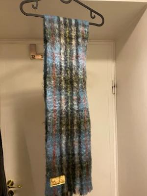 Rutig mohairhalsduk från Craig-Na-Creidhe - Säljer en fluffig halsduk från Craig-Na-Creidhe, tillverkad i Skottland. Västen har en mix av blå, grå, svart, grön och röda toner och är gjord av 75% mohair, 21% ull och 4% nylon. Perfekt för dig som gillar retro och statement-plagg.