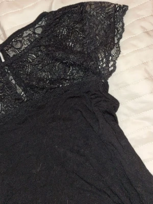 Svart blus med spetsdetaljer - Säljer en svart blus med vackra spetsdetaljer upptill och på ärmarna. Blusen har en feminin och elegant look med korta ärmar och rund hals. Materialet är mjukt och lätt, perfekt för dig som gillar stilrena plagg med lite extra detaljer.