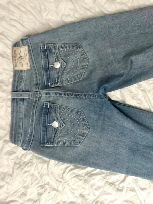 True Religion ljusblå jeans med ficklock - Säljer ett par ljusblå jeans från True Religion med ikoniska vita sömmar och stora ficklock bak med silverfärgade knappar. Klassisk femficksmodell, rak passform och snyggt broderad logga på bakfickorna. Perfekt för dig som gillar street och detaljer.
