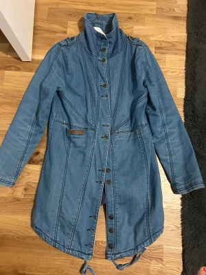 Blå jeansjacka från Red Issue - Snygg lång jeansjacka från Red Issue i blå denim med hög krage och coola detaljer som knappar framtill och snörning nedtill. Jackan har markerade sömmar och en liten läderpatch med logga vid fickan. Perfekt för dig som gillar street och vill sticka ut.
