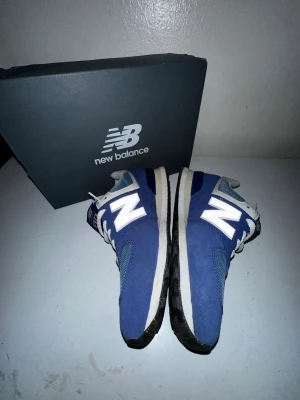 Blå New Balance 574 sneakers - Säljer ett par klassiska New Balance 574 sneakers i blått med vita detaljer och grå meshpartier. Skorna har en robust svart sula, vit N-logga på sidan och snörning. Perfekta för dig som gillar retrostil och bekväma sneakers. Storlek 44 (EU), 10 (US). Priset kan självklart diskuteras.