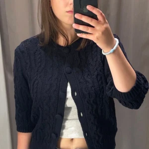 Mörkblå stickad kofta från Zara - Säljer en mörkblå stickad kofta från Zara med snyggt kabelstickat mönster och stora matchande knappar framtill. Koftan har halvlånga ärmar och ribbade muddar vid ärmslut och nederkant. Sitter som en S