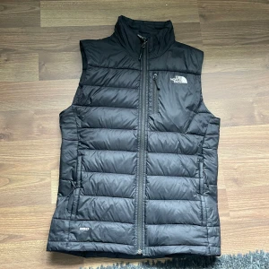 Svart dunväst från The North Face - En svart dunväst från The North Face med dragkedja framtill och en bröstficka med dragkedja. Västen är quiltad och har en hög krage för extra värme. inga jättesynliga tecken på användning 