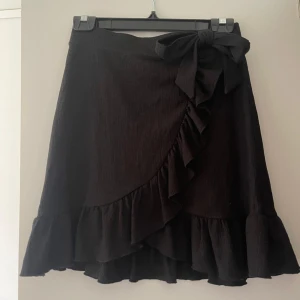Svart kjol med volang och rosett - Säljer denna eleganta svarta kjolen med volangdetaljer och knyte på sidan. Kjolen har en omlott-design och är kort i längden. Köpt på Ginatricot och endast använd en gång. ✨🤍🖤