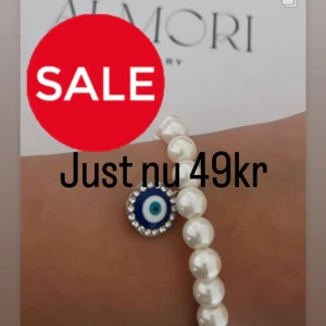 Pärlarmband med blått öga - Ett elegant pärlarmband med vita pärlor och ett blått öga som hänge. Hänget är dekorerat med små glittriga stenar runt om.