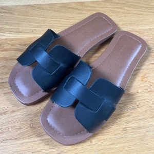 Bruna sandaler med svart rem - Ett par bruna sandaler med en bred svart rem över foten. Sandalerna har en enkel och stilren design med en fyrkantig tåform.