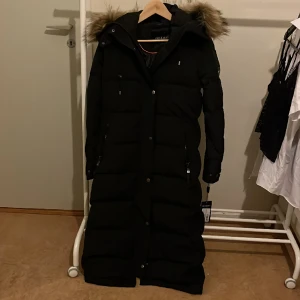 Hollies Metro Dunjacka - Nyskick dunjacka från Hollies. Nypris 6249 kr, säljer för 4500 kr. Dunjackan har en formsydd, smickrande siluett och en dekorativ knapprad mitt fram. En längre modell som verkligen håller dig varm under vintern.