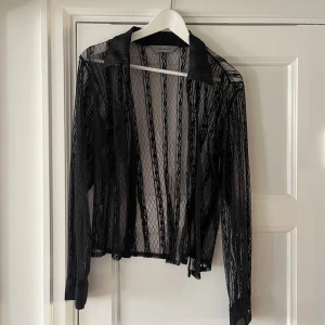 Svart spetsjacka - En elegant svart spetsskjorta med långa ärmar och krage. Skjortan är genomskinlig och har ett detaljerat spetsmönster. Den har knappar framtill och små stenar i svart!