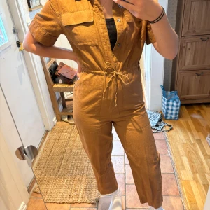 Weekday brun jumpsuit - En snygg jumpsuit i en härlig brun färg. Den har en skön passform och är perfekt för både vardag och fest. Jumpsuiten har en dragkedja framtill och en dragsko i midjan. Den har även två fickor framtill!