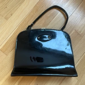 Vintage svart handväska med metallknäppning - Vintage svart högblank handväska. Snygg metallknäppning i guldig färg. Lite smårepor men inget som stör. Väskan är ca 22x25x7 + handtag. 